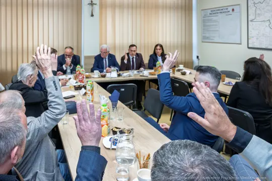 Spotkanie grupy ludzi przy stole, podnoszących ręce w głosowaniu.