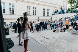 Prezentacja przed dużą grupą ludzi na zewnątrz, w tle budynki i krzesła.