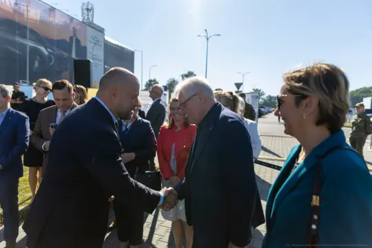 Spotkanie dwóch mężczyzn na tle grupy osób, w tym kobiet, w słoneczny dzień.