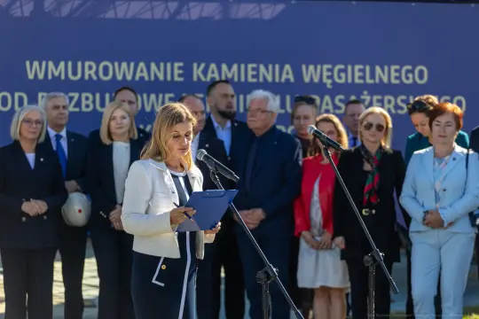Kobieta przemawia na tle grupy ludzi podczas uroczystości wmurowania kamienia węgielnego.