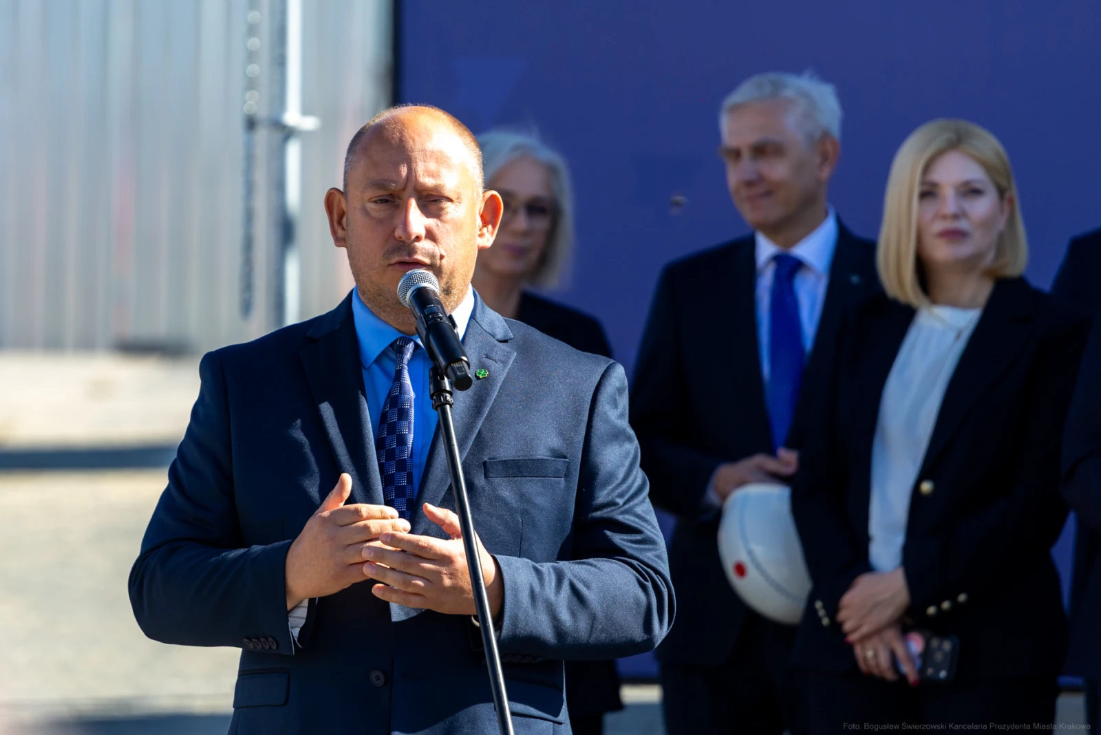 Mężczyzna w garniturze przemawia do mikrofonu, w tle grupa osób.  Autor: Bogusław Świerzowski