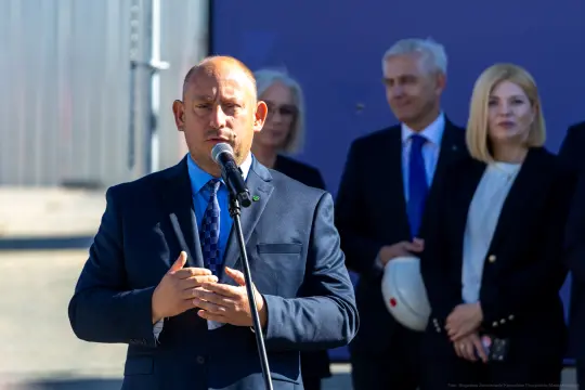 Mężczyzna w garniturze przemawia do mikrofonu, w tle grupa osób.
