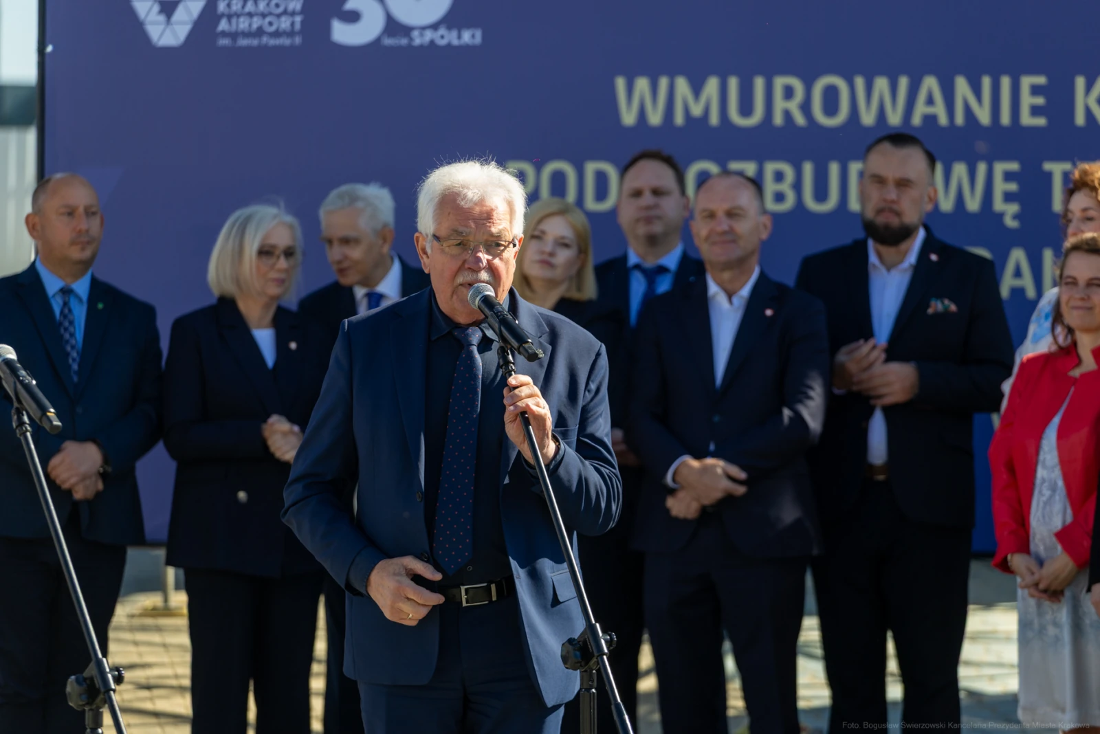 Mężczyzna w garniturze przemawia do mikrofonu, w tle grupa osób i baner z napisem.  Autor: Bogusław Świerzowski