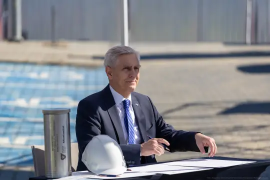 Mężczyzna w garniturze siedzi przy stole, podpisując dokumenty, obok kask budowlany.