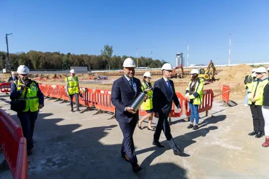 Uczestnicy ceremonii otwarcia budowy, w kaskach i kamizelkach odblaskowych, trzymający elementy budowlane.