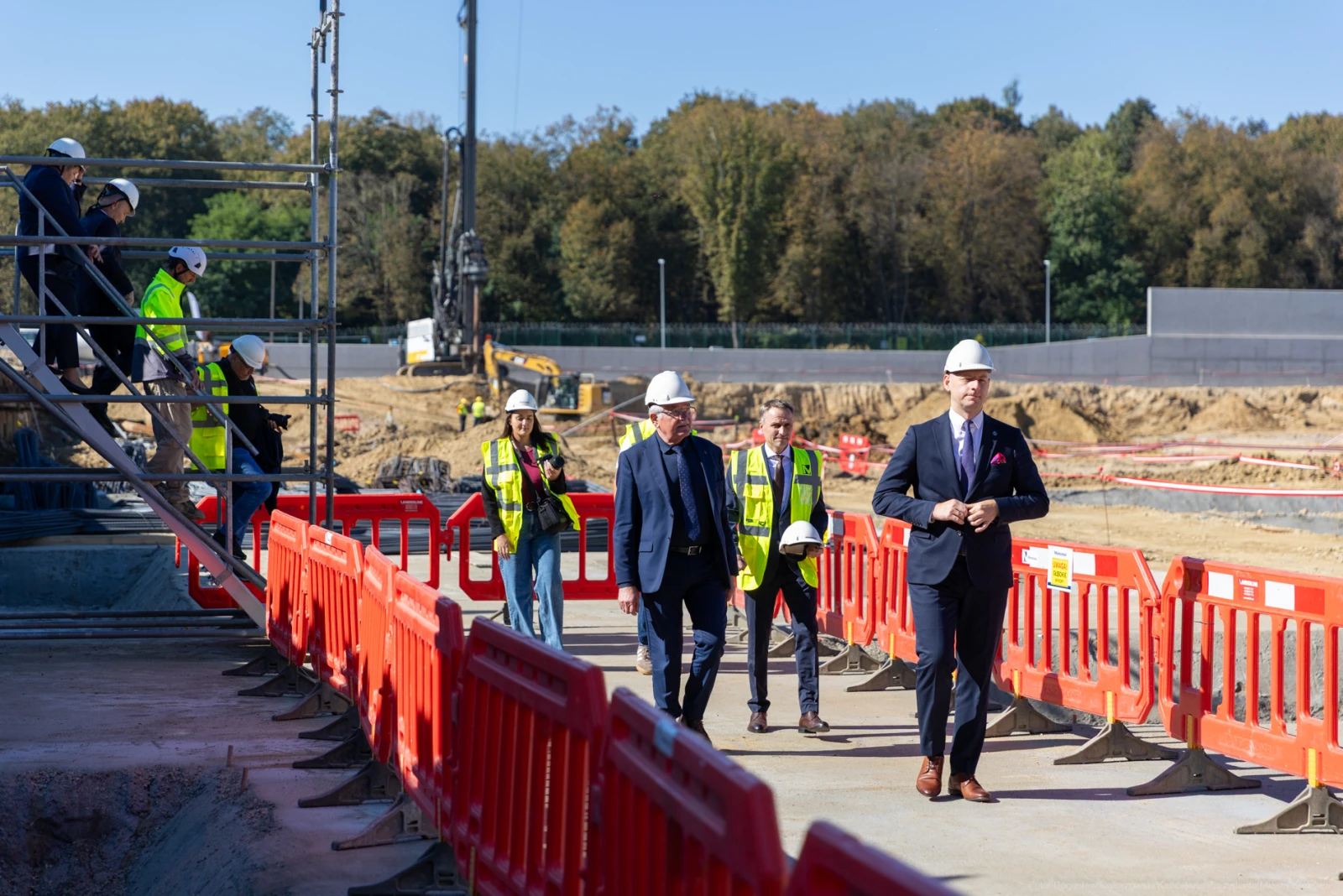 Grupa ludzi w kaskach na placu budowy, w tle maszyny budowlane i ogrodzenia.  Autor: Bogusław Świerzowski