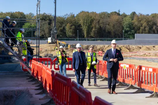 Grupa ludzi w kaskach na placu budowy, w tle maszyny budowlane i ogrodzenia.