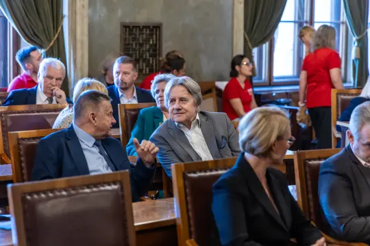 Grupa osób w sali, niektórzy rozmawiają, inni siedzą w ciszy.