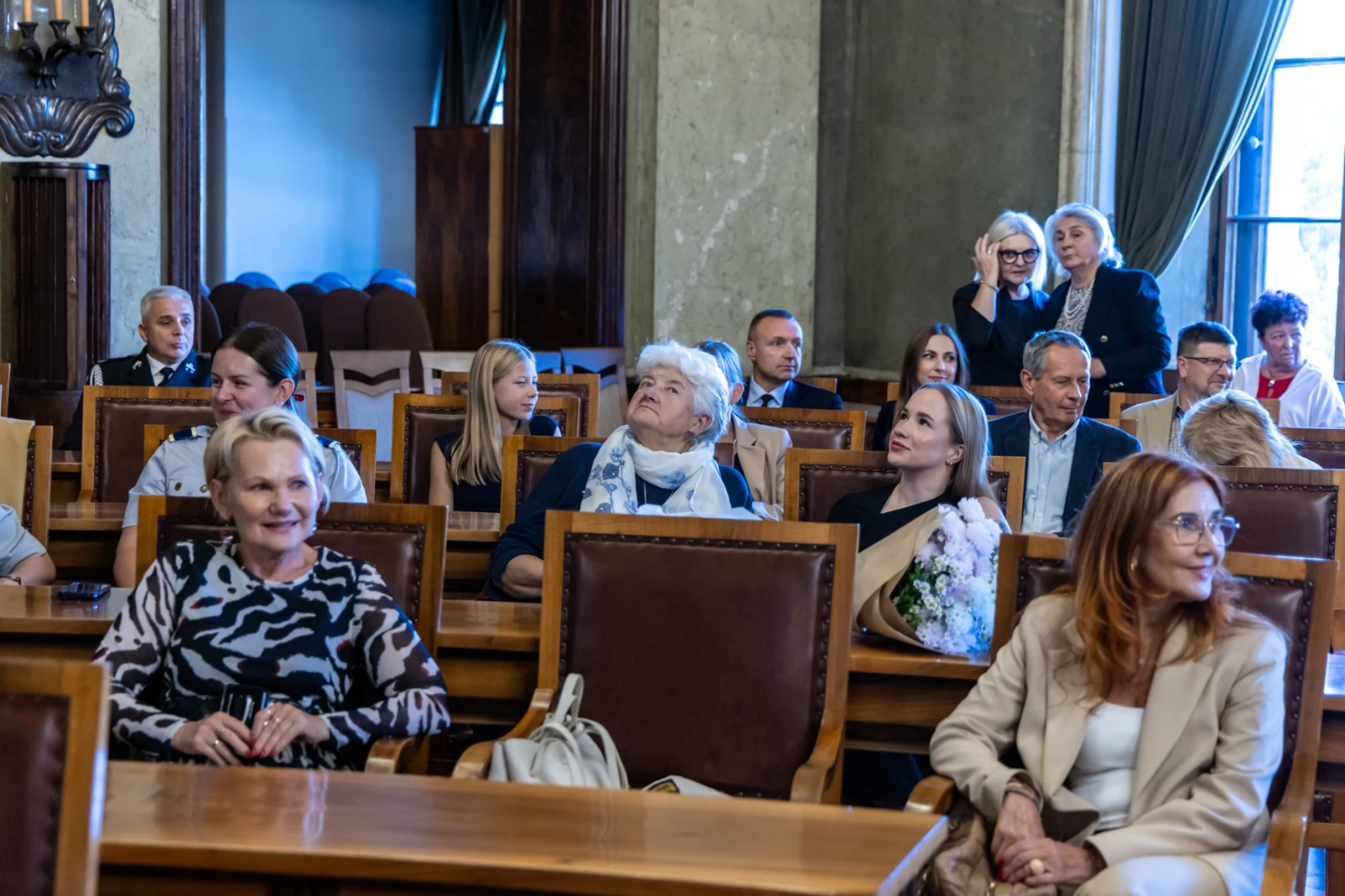 Grupa osób siedzących w sali, niektórzy z bukietami kwiatów.  Autor: Jan Graczyński
