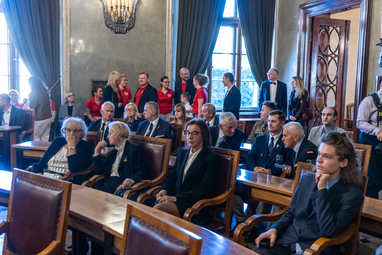 Grupa ludzi w sali, część w garniturach, część w strojach formalnych, siedzących przy stołach.  Autor: Jan Graczyński