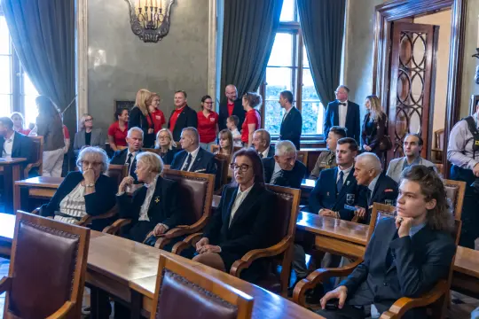 Grupa ludzi w sali, część w garniturach, część w strojach formalnych, siedzących przy stołach.