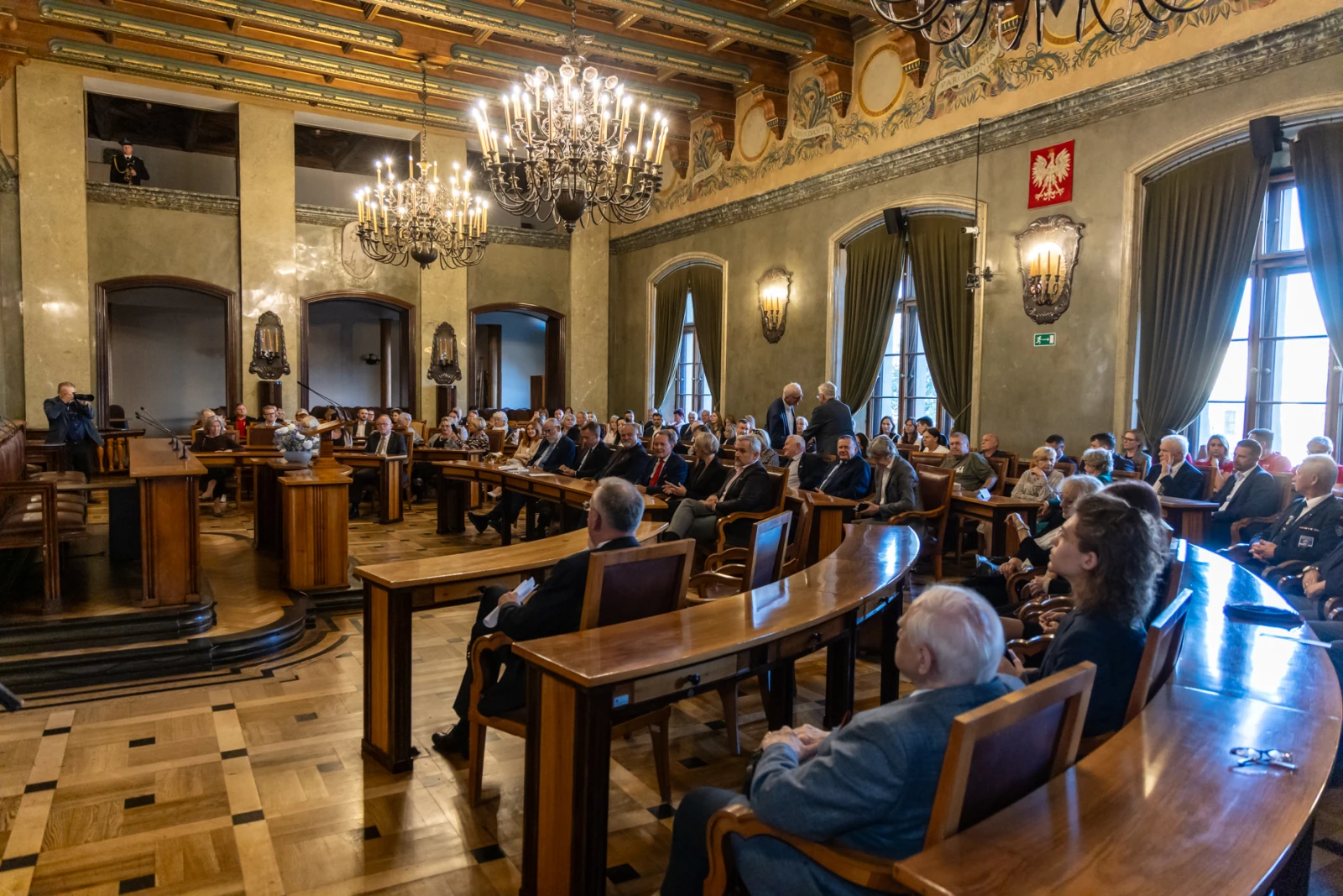 Sala z dużą ilością osób siedzących w rzędach, eleganckie wnętrze z żyrandolami.  Autor: Jan Graczyński