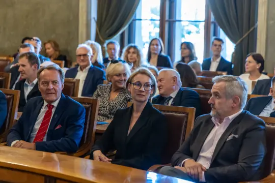 Grupa osób siedzących w sali konferencyjnej, wśród nich kobieta w okularach i mężczyźni w garniturach.