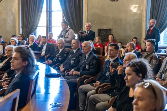 Grupa ludzi w sali obrad, niektórzy w mundurach, inni w formalnych strojach.