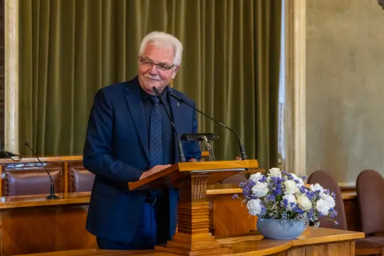 Mężczyzna w garniturze przemawia przy mównicy, obok bukiet kwiatów.