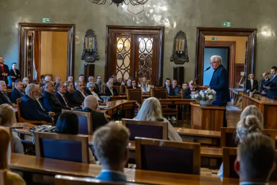 Uroczystość w sali obrad z przemawiającym mężczyzną i zebranymi gośćmi.