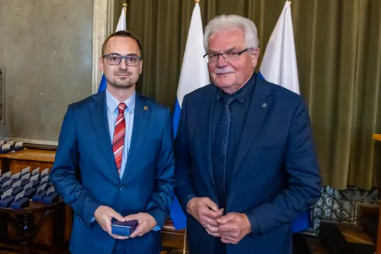 Dwóch mężczyzn w garniturach, jeden z medalem, stojących obok siebie w sali z flagami.