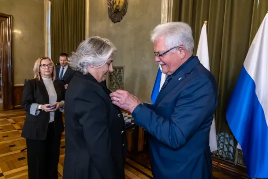Wręczenie odznaczenia przez mężczyznę kobiecie w formalnym otoczeniu.