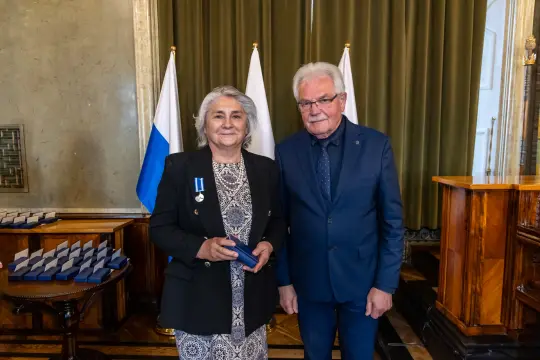 Wręczenie odznaczenia kobiecie w towarzystwie mężczyzny, w tle flagi i stoły z nagrodami.