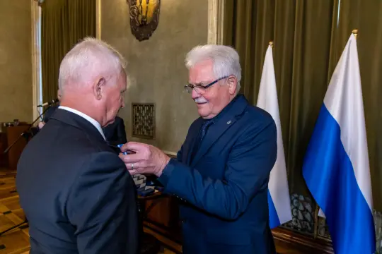 Wręczenie odznaczenia przez mężczyznę w garniturze drugiemu mężczyźnie. W tle flagi.