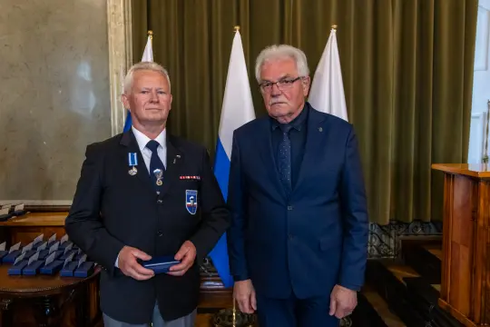 Dwaj mężczyźni w garniturach, jeden z odznaczeniami, stoją w sali z flagami.