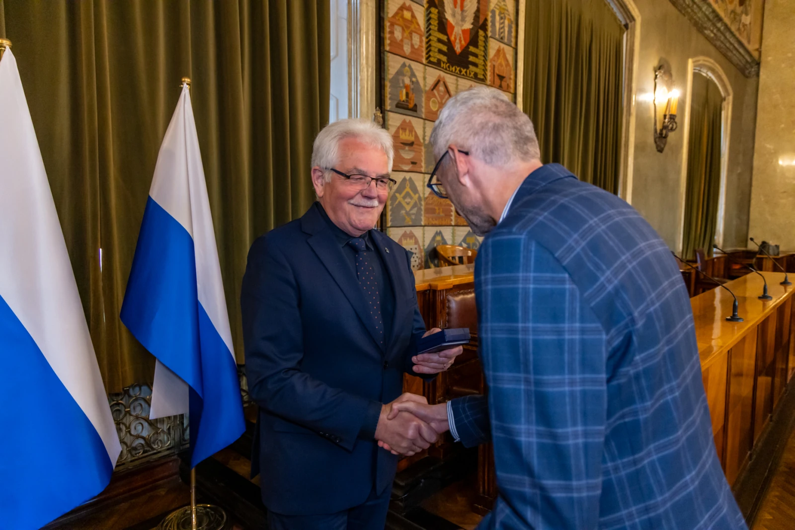 Mężczyzna w garniturze uścisnął dłoń innemu mężczyźnie w formalnym otoczeniu. Flagi w tle.  Autor: Jan Graczyński