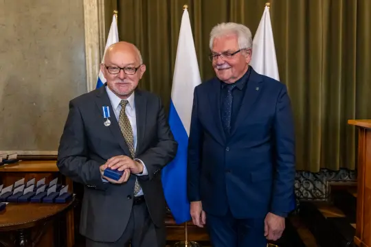 Dwaj mężczyźni w garniturach, jeden z medalem, stoją obok flagi.