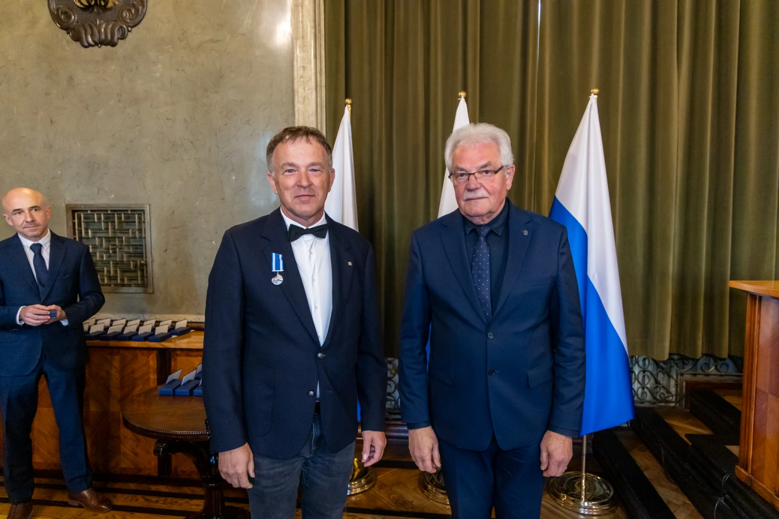 Dwóch mężczyzn w garniturach, jeden z medalem, stojących obok flagi.  Autor: Jan Graczyński
