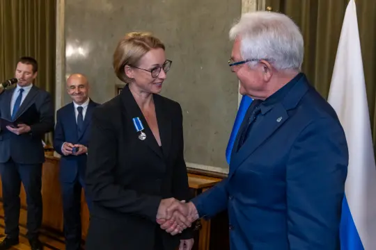 Wręczenie odznaczenia, uściski dłoni, uśmiechnięte twarze, oficjalna ceremonia.