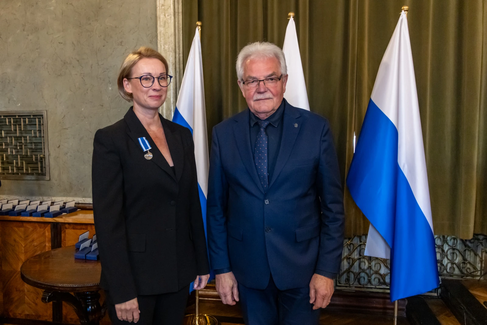 Wręczenie odznaczenia, dwie osoby w formalnych strojach, flagi w tle.  Autor: Jan Graczyński