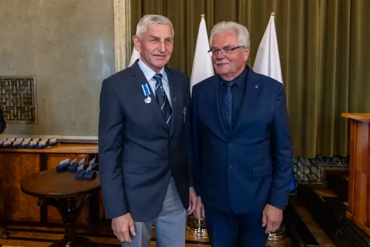 Dwaj mężczyźni w garniturach, jeden z medalem, stoją obok siebie w eleganckim wnętrzu.