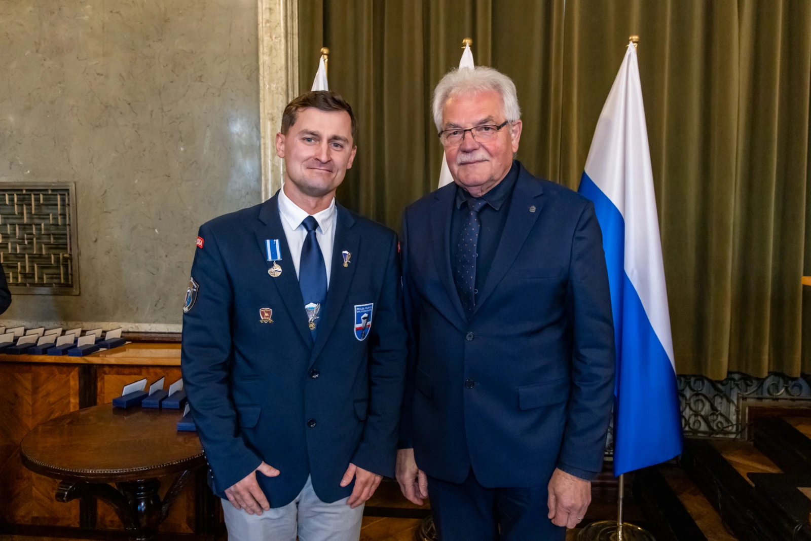 Mężczyźni w garniturach, jeden z medalami, stoją obok flagi.  Autor: Jan Graczyński