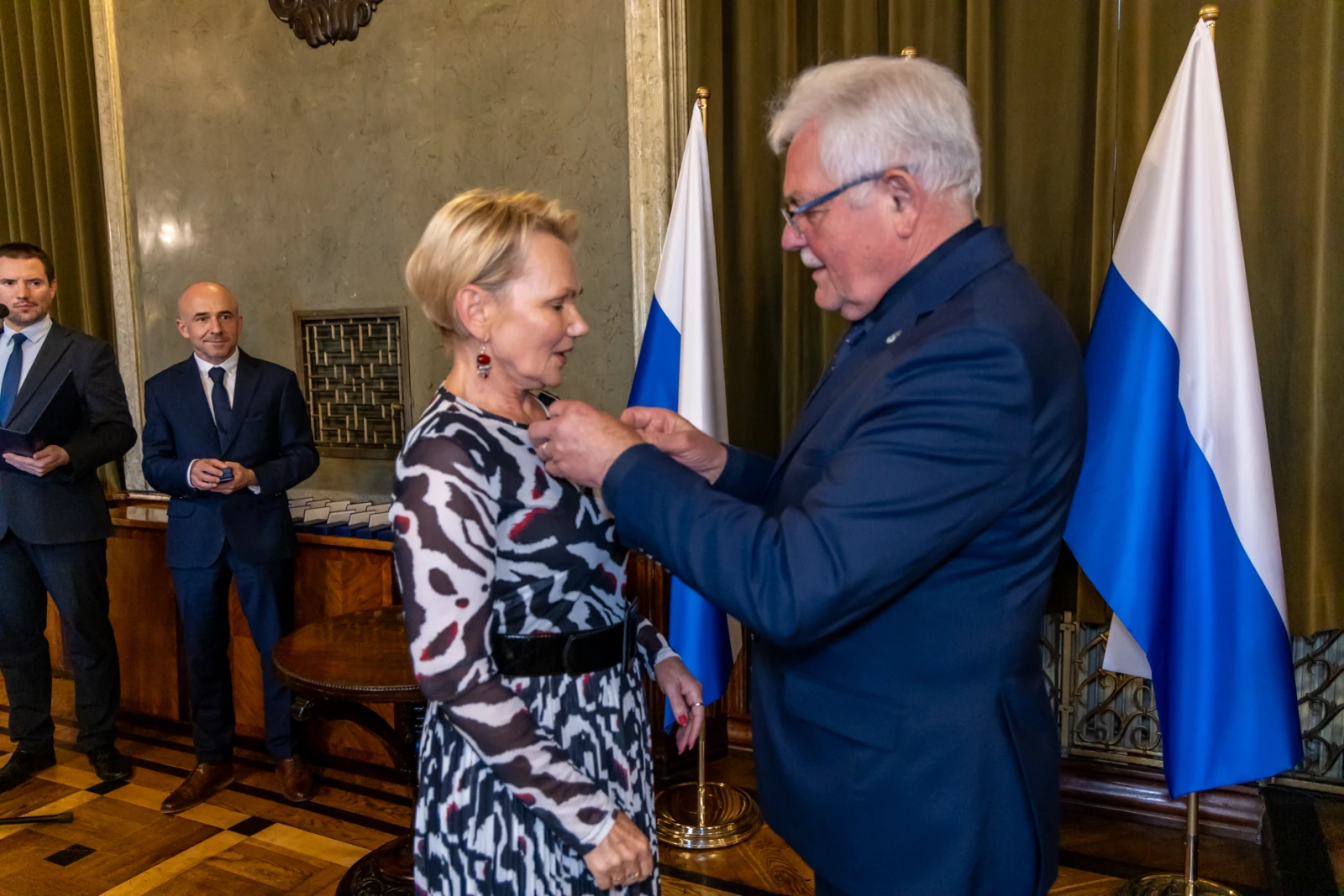 Wręczenie odznaczenia kobiecie przez mężczyznę w eleganckim otoczeniu.  Autor: Jan Graczyński