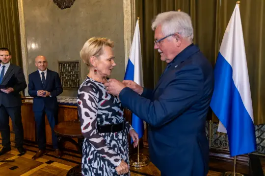 Wręczenie odznaczenia kobiecie przez mężczyznę w eleganckim otoczeniu.