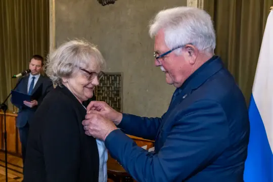 Wręczenie odznaczenia kobiecie przez mężczyznę w oficjalnym otoczeniu.