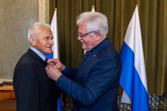 Dwaj mężczyźni w garniturach, jeden przypina odznaczenie drugiemu, w tle flagi.