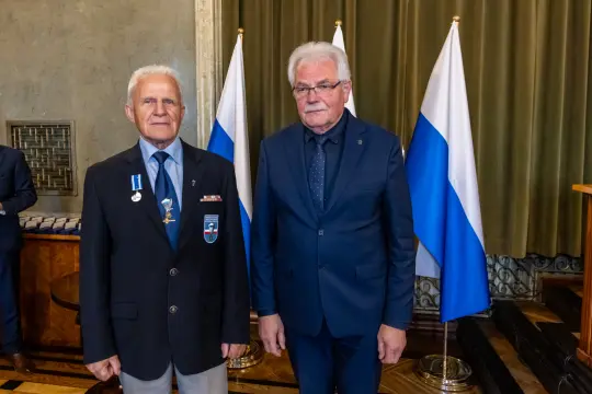 Dwaj mężczyźni w garniturach, jeden z odznaczeniem, stoją obok siebie w formalnym otoczeniu.