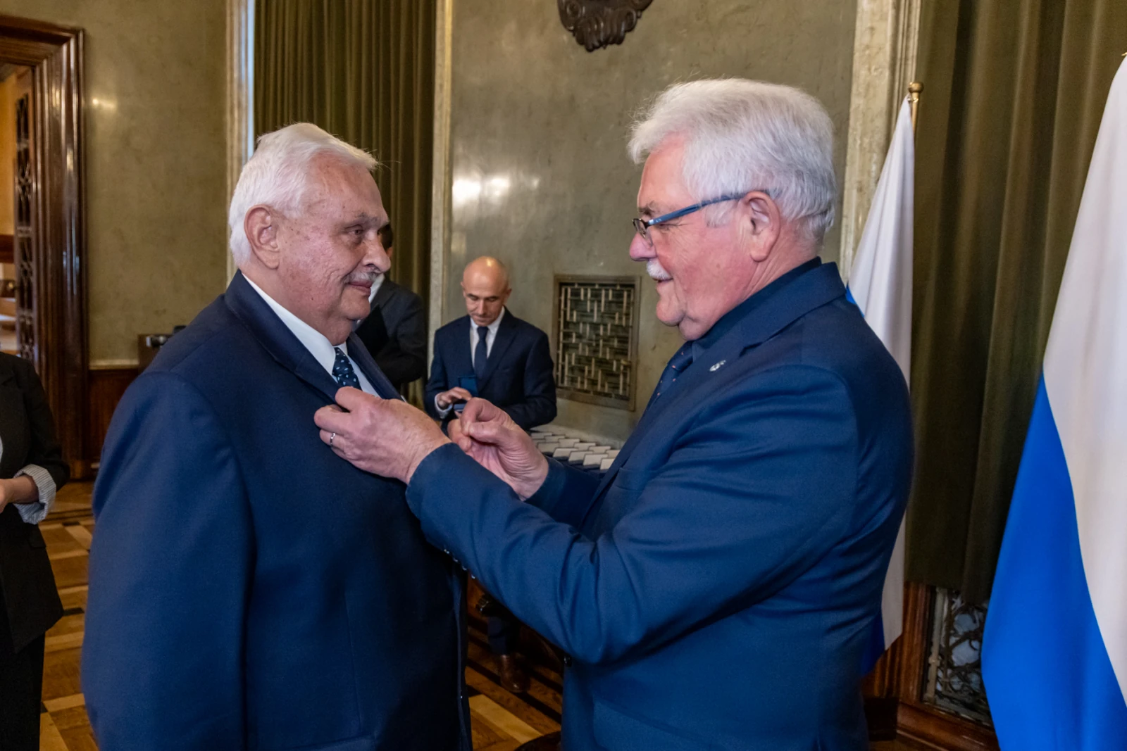 Wręczenie odznaczenia mężczyźnie w garniturze przez innego mężczyznę w formalnym ubraniu.  Autor: Jan Graczyński
