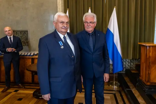 Dwaj mężczyźni w garniturach, jeden z medalem, stoją w sali z flagami.