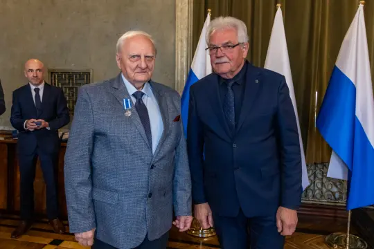 Dwaj mężczyźni w garniturach, jeden z medalem, stoją obok siebie w sali z flagami.
