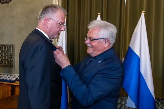 Mężczyzna przypina odznaczenie do marynarki innego mężczyzny w eleganckim otoczeniu.