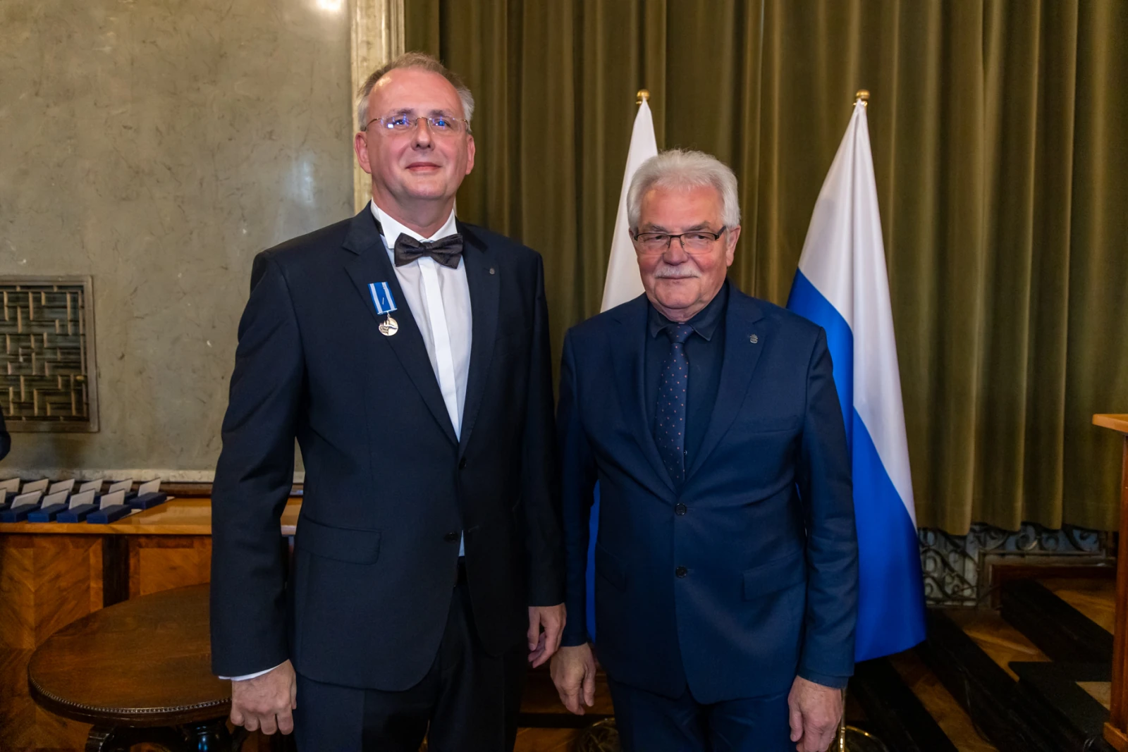 Dwaj mężczyźni w eleganckich strojach, jeden z medalem, stoją obok siebie w formalnym otoczeniu.  Autor: Jan Graczyński