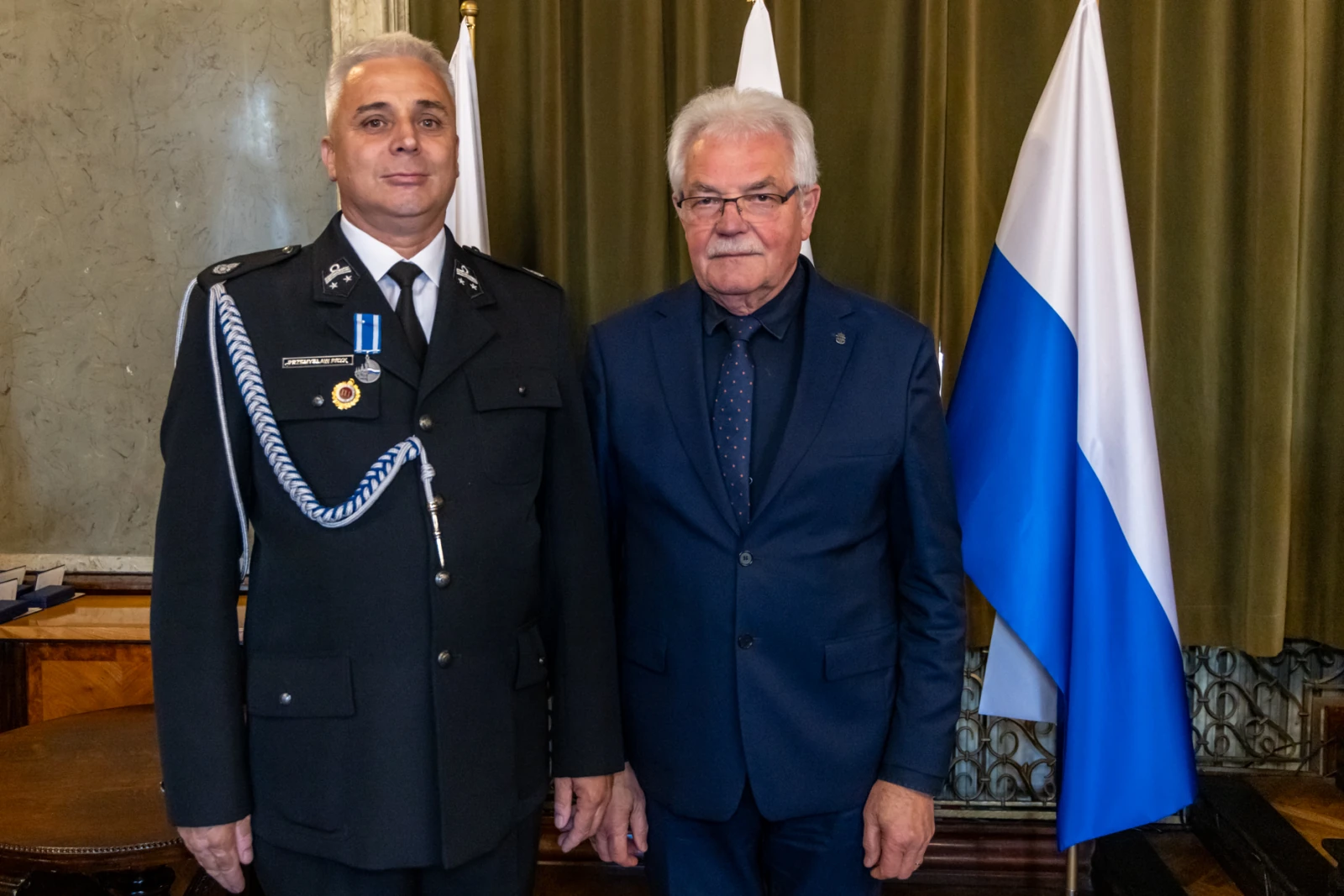 Dwaj mężczyźni w garniturach, jeden w mundurze, stoją obok siebie, za nimi flagi.  Autor: Jan Graczyński