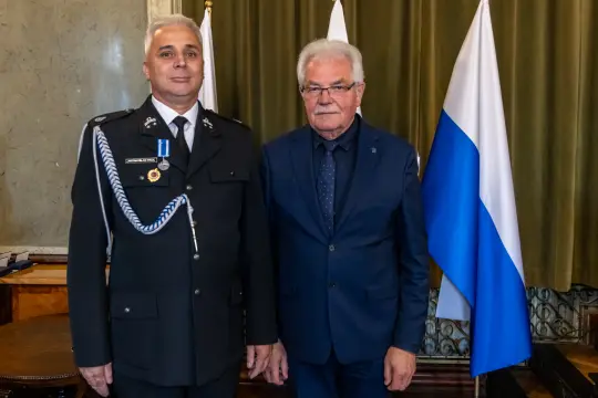 Dwaj mężczyźni w garniturach, jeden w mundurze, stoją obok siebie, za nimi flagi.