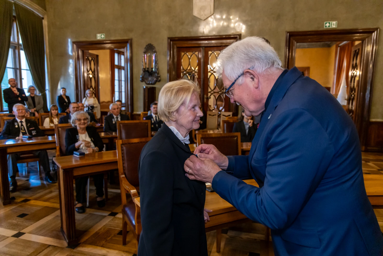 Wręczenie odznaczenia kobiecie w eleganckim otoczeniu, z obecnością gości.  Autor: Jan Graczyński
