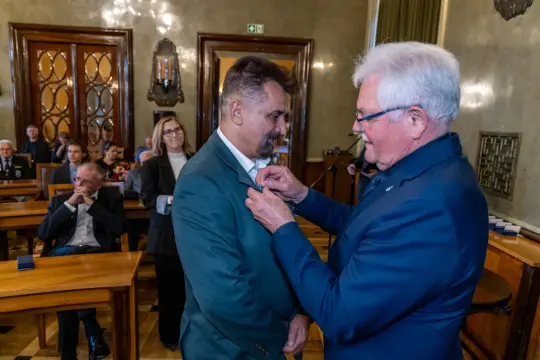 Mężczyzna otrzymuje odznaczenie w sali z drewnianymi meblami.