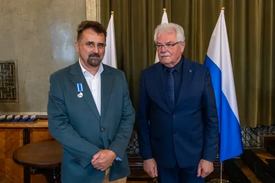Dwaj mężczyźni w garniturach, jeden z odznaczeniem, stoją obok flag.