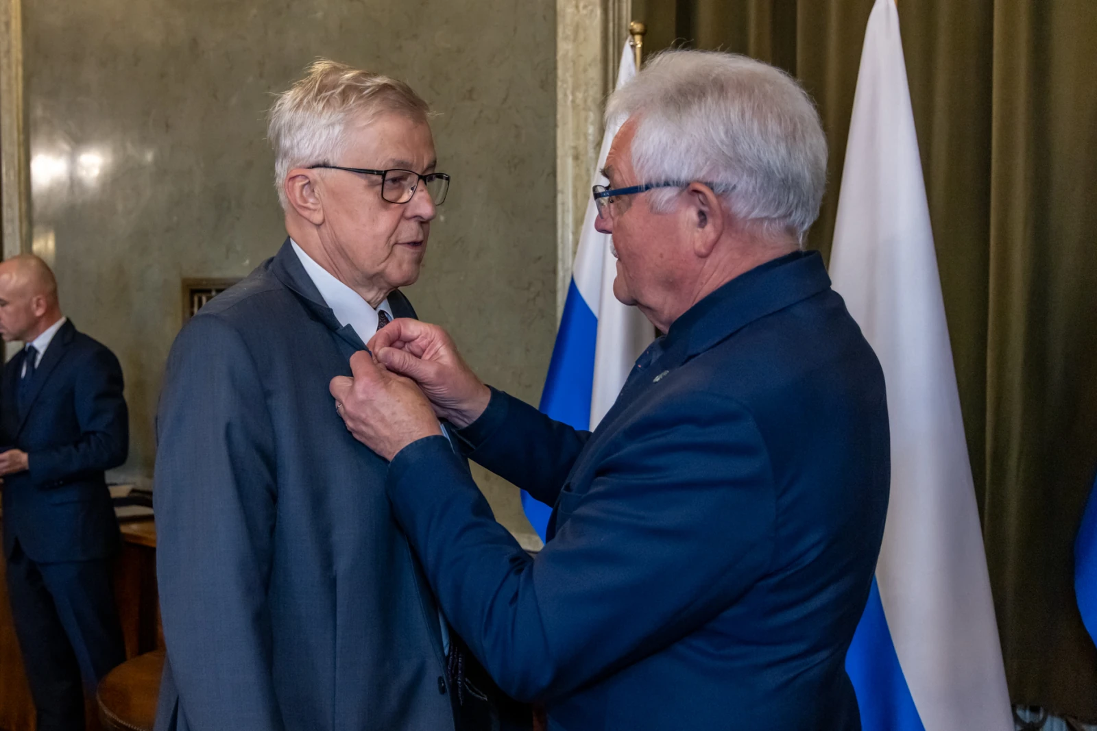 Mężczyzna odbiera odznaczenie w obecności innych osób i flag.  Autor: Jan Graczyński
