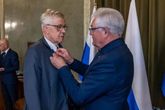 Mężczyzna odbiera odznaczenie w obecności innych osób i flag.