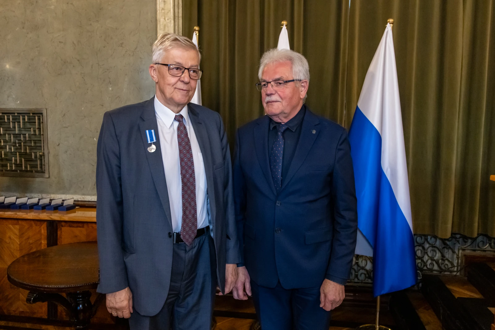 Dwaj mężczyźni w garniturach, jeden z odznaczeniem, stoją obok siebie w eleganckim wnętrzu.  Autor: Jan Graczyński